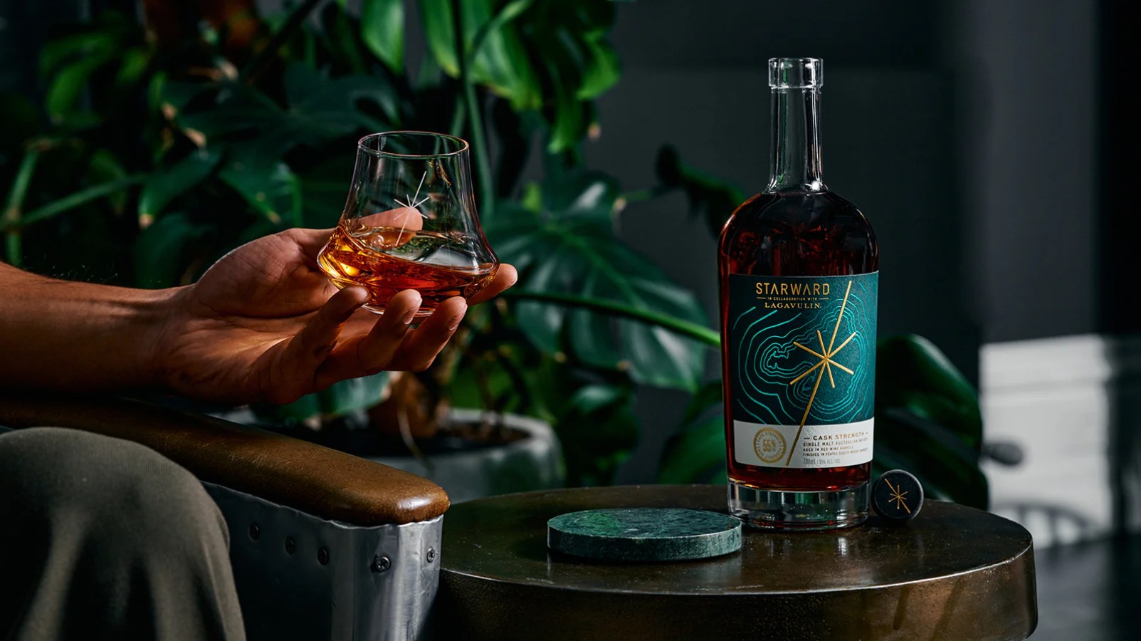 A hand holding a whiskey glass beside a bottle of Starward cask strength, made in collaboration with Lagavulin